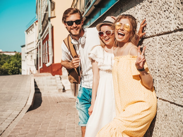 Grupo de jóvenes tres elegantes amigos posando en la calle. Hombre de moda y dos chicas lindas vestidas con ropa casual de verano. Modelos sonrientes divirtiéndose en gafas de sol. Mujeres alegres y chico al aire libre