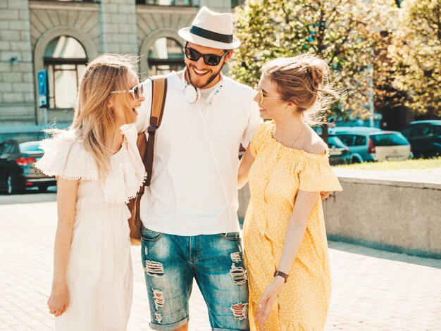 Grupo de jóvenes tres elegantes amigos posando en la calle. Hombre de moda y dos chicas lindas vestidas con ropa casual de verano. Modelos sonrientes divirtiéndose en gafas de sol. Mujeres alegres y chico chateando