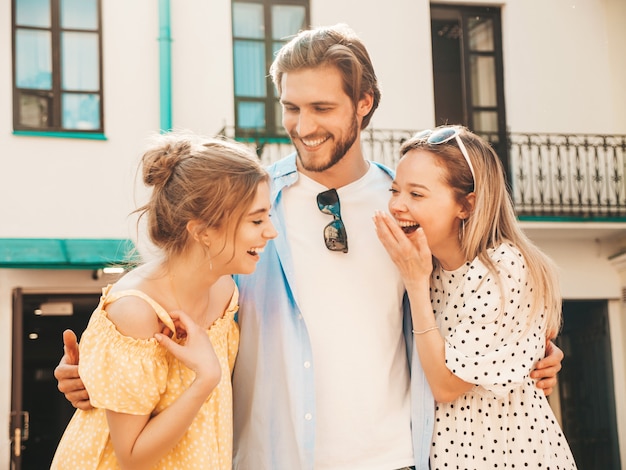 Grupo de jóvenes tres elegantes amigos posando en la calle. Hombre de moda y dos chicas lindas vestidas con ropa casual de verano. Modelos sonrientes divirtiéndose en gafas de sol. Mujeres alegres y chico chateando