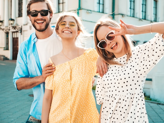 Grupo de jóvenes tres elegantes amigos posando en la calle. Hombre de moda y dos chicas lindas vestidas con ropa casual de verano. Modelos sonrientes divirtiéndose en gafas de sol. Mujeres alegres y chico en susnet