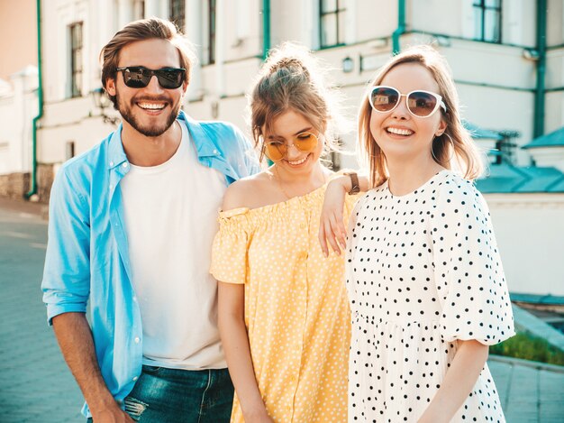 Grupo de jóvenes tres elegantes amigos posando en la calle. Hombre de moda y dos chicas lindas vestidas con ropa casual de verano. Modelos sonrientes divirtiéndose en gafas de sol. Mujeres alegres y chico en susnet