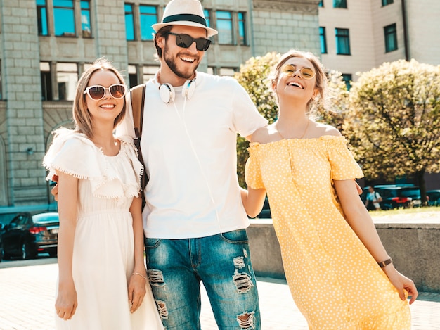 Foto gratuita grupo de jóvenes tres elegantes amigos posando en la calle. hombre de moda y dos chicas lindas vestidas con ropa casual de verano. modelos sonrientes divirtiéndose en gafas de sol. mujeres alegres y un chico volviéndose loco