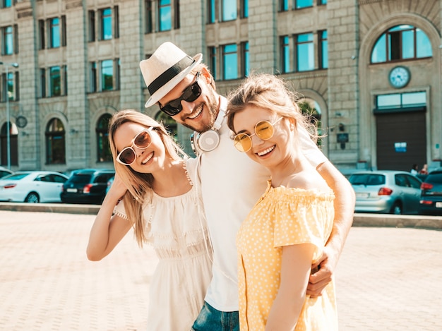 Grupo de jóvenes tres elegantes amigos posando en la calle. Hombre de moda y dos chicas lindas vestidas con ropa casual de verano. Modelos sonrientes divirtiéndose en gafas de sol. Mujeres alegres y un chico volviéndose loco