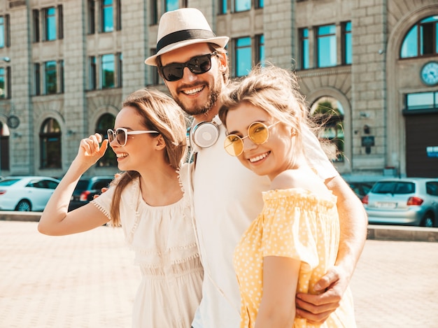 Grupo de jóvenes tres elegantes amigos posando en la calle. Hombre de moda y dos chicas lindas vestidas con ropa casual de verano. Modelos sonrientes divirtiéndose en gafas de sol. Mujeres alegres y un chico volviéndose loco