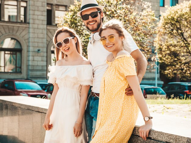 Grupo de jóvenes tres elegantes amigos posando en la calle. Hombre de moda y dos chicas lindas vestidas con ropa casual de verano. Modelos sonrientes divirtiéndose en gafas de sol. Mujeres alegres y un chico volviéndose loco