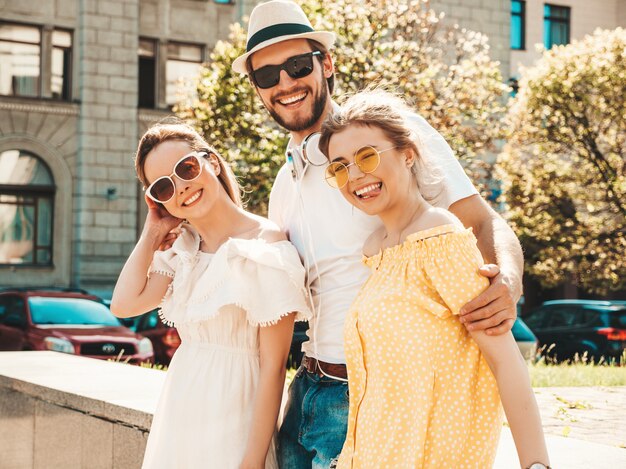 Grupo de jóvenes tres elegantes amigos posando en la calle. Hombre de moda y dos chicas lindas vestidas con ropa casual de verano. Modelos sonrientes divirtiéndose en gafas de sol. Mujeres alegres y un chico volviéndose loco