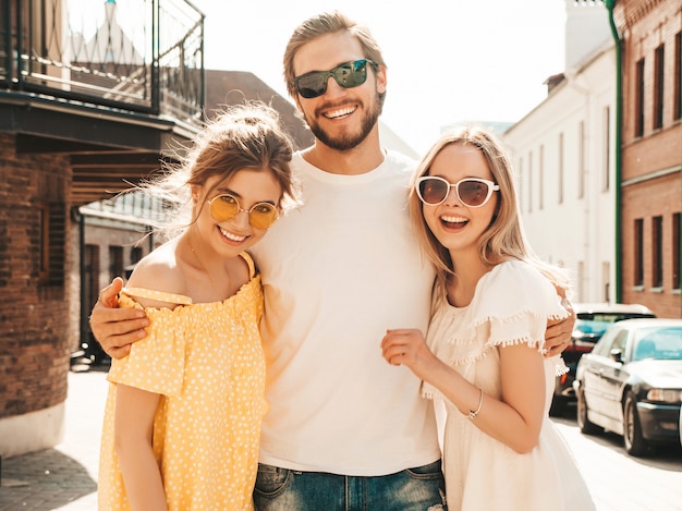 Grupo de jóvenes tres elegantes amigos posando en la calle. Hombre de moda y dos chicas lindas vestidas con ropa casual de verano. Modelos sonrientes divirtiéndose en gafas de sol. Mujeres alegres y un chico volviéndose loco