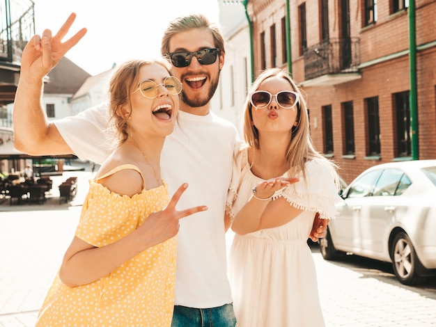 Grupo de jóvenes tres elegantes amigos posando en la calle. Hombre de moda y dos chicas lindas vestidas con ropa casual de verano. Modelos sonrientes divirtiéndose en gafas de sol. Mujeres alegres y un chico volviéndose loco