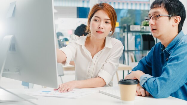 Grupo milenario de joven empresario y empresaria de Asia en la pequeña oficina urbana moderna. Supervisor de jefe masculino japonés enseñando pasante o nueva empleada coreana ayudando con tareas difíciles.