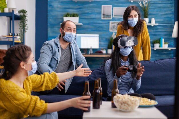 Grupo mixto de personas guiando a una mujer negra con auriculares vr jugando videojuegos virtuales en la sala de estar manteniendo el distanciamiento social contra el covid19. Diversos amigos divirtiéndose en una nueva fiesta normal.