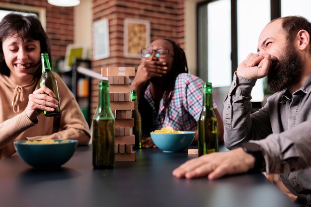 Grupo multicultural de amigos riéndose de todo corazón mientras juegan juegos de sociedad juntos en casa. Felices y alegres personas diversas sentadas a la mesa en la sala de estar mientras juegan con bloques de madera.