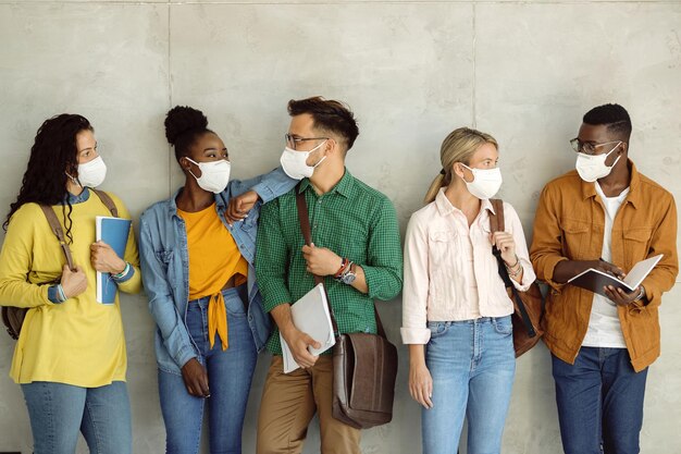 Grupo multiétnico de amigos universitarios que se comunican mientras usan máscaras protectoras mientras están de pie contra la pared en un vestíbulo.