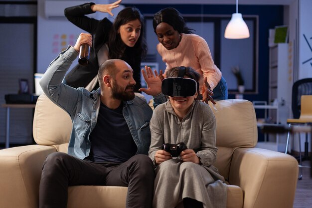 Grupo multiétnico de colegas jugando videojuegos con gafas vr en la fiesta de la oficina después de las horas de trabajo. Mujer triste perdiendo el juego en la consola con auriculares y controlador de realidad virtual.