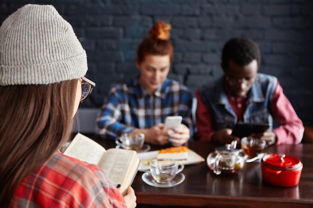 Foto gratuita grupo multiétnico de elegantes jóvenes estudiantes bebiendo té en el café durante las vacaciones: mujer con sombrero leyendo un libro mientras pelirroja mujer y hombre africano con aparatos electrónicos.