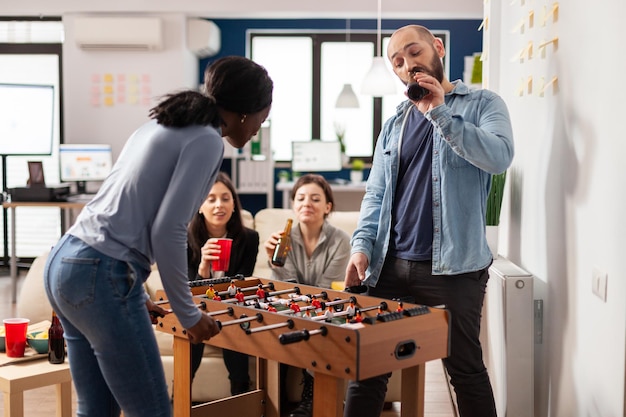 Grupo multiétnico de personas jugando en el juego de mesa de futbolín, reuniéndose para tomar una copa en la oficina después de las horas de trabajo. Hombre y mujer bebiendo cerveza de botellas y comiendo bocadillos en la celebración.