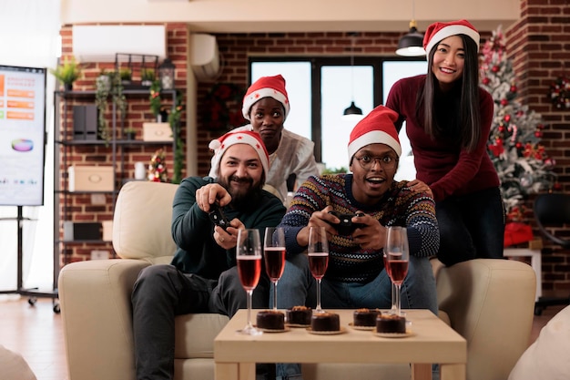 Grupo multiétnico de personas jugando videojuegos, usando sombrero de santa en la fiesta festiva de la oficina. Celebrando la festividad de la víspera de navidad en el lugar de trabajo con decoraciones navideñas, divirtiéndose con la consola de juegos.