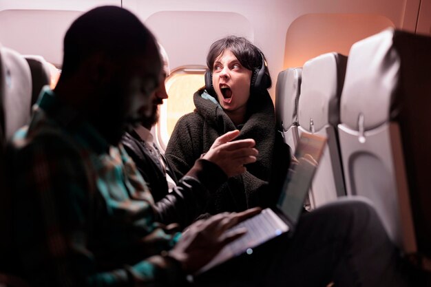 Grupo multiétnico de personas que vuelan al extranjero en clase económica, usando laptop, smartphone y auriculares durante el vuelo al atardecer. Pasajeros que viajan con líneas aéreas comerciales internacionales.