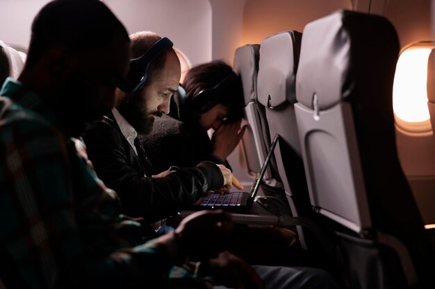 Grupo multiétnico de personas que vuelan juntas en clase económica para llegar a destino, hombre que usa computadora portátil durante un vuelo internacional. Turistas que viajan al extranjero en avión.