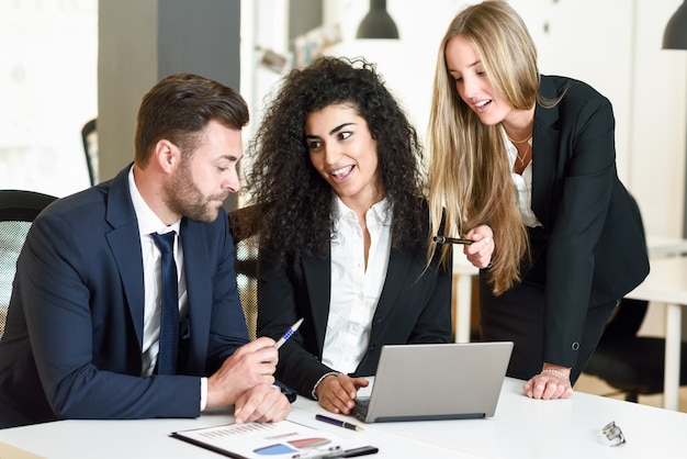 Grupo multiétnico de tres empresarios reunión en una oficina moderna. Dos mujeres y un hombre caucásico llevaba traje mirando un ordenador portátil.