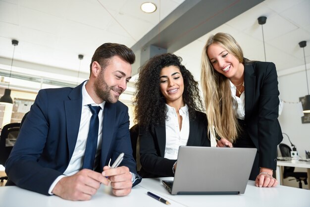 Grupo multiétnico de tres empresarios reunión en una oficina moderna. Dos mujeres y un hombre llevando traje mirando un ordenador portátil.