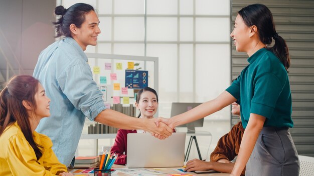 Grupo multirracial de jóvenes creativos en ropa casual elegante discutiendo negocios dándose la mano y sonriendo mientras está de pie en la oficina moderna. Cooperación de socios, concepto de trabajo en equipo de compañeros de trabajo.