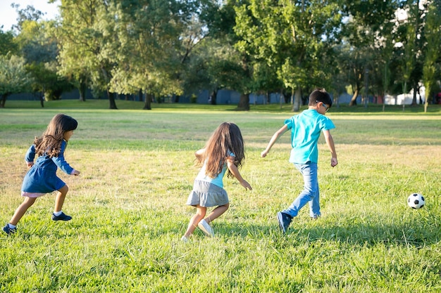 Grupo de niños activos jugando al fútbol en el césped en el parque de la ciudad. Vista trasera completa. Concepto de actividad infantil y al aire libre.