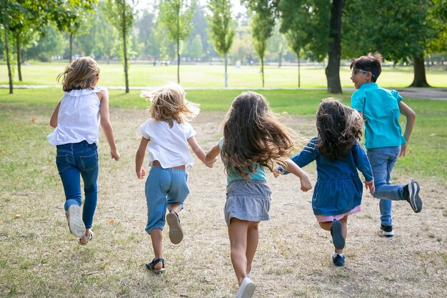 Grupo de niños corriendo juntos sobre la hierba, con carrera en el parque. Vista posterior, de cuerpo entero. Concepto de actividad al aire libre para niños