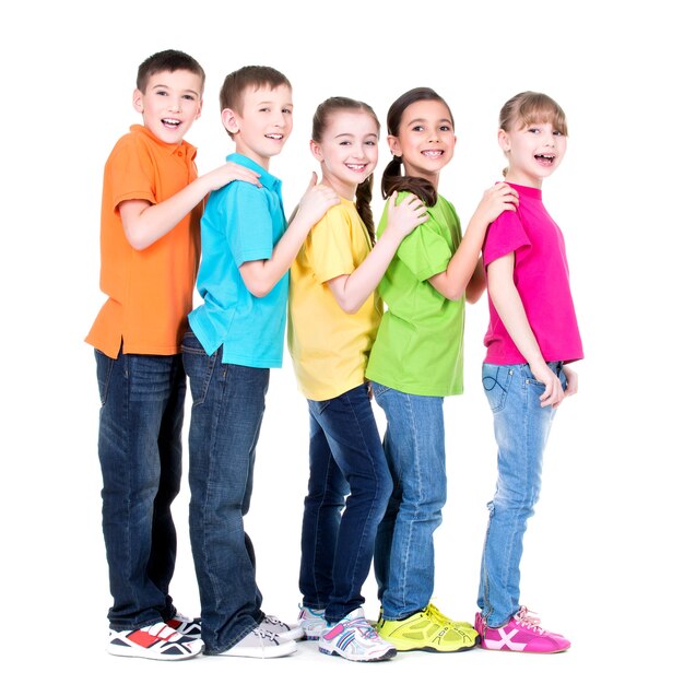 Grupo de niños felices en camisetas de colores se colocan uno detrás del otro poniendo las manos sobre los hombros sobre fondo blanco.