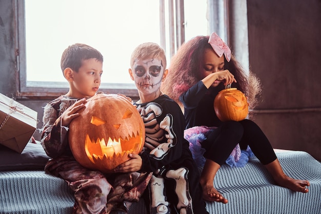 Un grupo de niños multirraciales durante la fiesta de Halloween sostiene calabazas mientras se sientan en la cama en una casa antigua. concepto de Halloween.