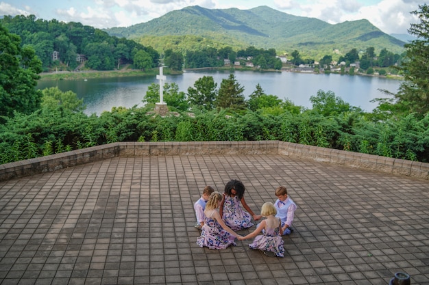 Foto gratuita grupo de niños rezando cerca de una cruz rodeada por un lago y colinas cubiertas de bosques