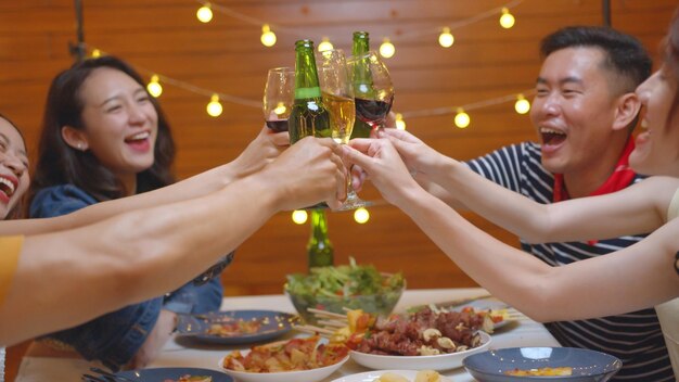 Grupo de personas asiáticas bebiendo alcohol animando una botella de cerveza y comiendo comida sentada en la mesa