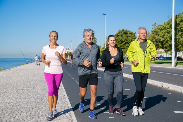 Grupo de personas maduras con ropa deportiva, trotar a lo largo de la orilla del río. Disparo de cuerpo entero. Concepto de jubilación o estilo de vida activo