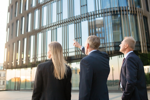 Grupo de socios comerciales en trajes formales apuntando al edificio de oficinas, reuniéndose al aire libre, discutiendo bienes raíces. Vista trasera. Concepto de bienes raíces comerciales