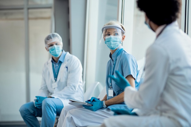 Grupo de trabajadores de la salud con ropa de trabajo protectora hablando mientras están sentados en un pasillo en una clínica médica