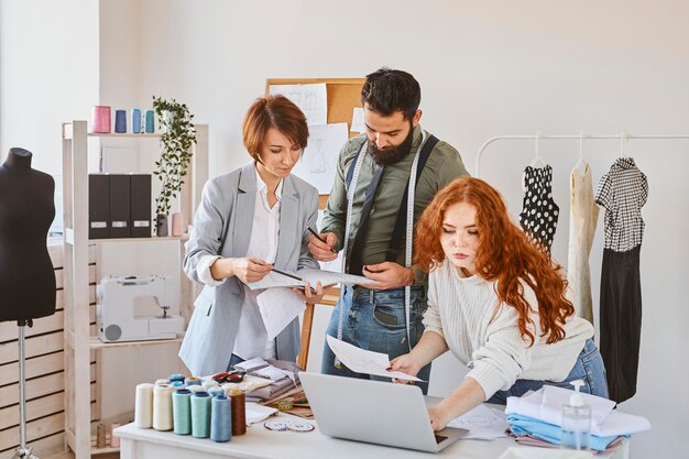 Grupo de tres diseñadores de moda que trabajan en el atelier con laptop y papeles