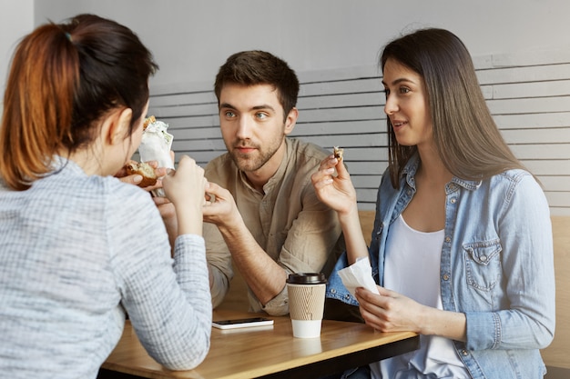 Grupo de tres guapos estudiantes sentados en la cafetería de la universidad, almorzando, hablando de los exámenes de ayer. Vida universitaria.