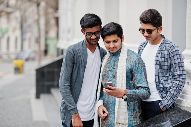 Grupo de tres hombres indios del sur de Asia con ropa tradicional e informal mirando el teléfono móvil