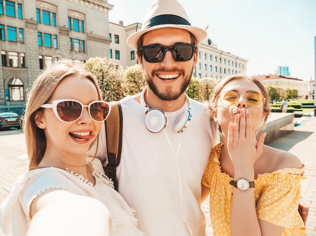 Grupo de tres jóvenes amigos con estilo en la calle. hombre y dos chicas lindas vestidas con ropa casual de verano. modelos sonrientes divirtiéndose en gafas de sol. mujeres y hombres haciendo fotos selfie en teléfono inteligente