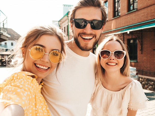 Grupo de tres jóvenes amigos con estilo en la calle. Hombre y dos chicas lindas vestidas con ropa casual de verano. Modelos sonrientes divirtiéndose en gafas de sol. Mujeres y hombres haciendo fotos selfie en teléfono inteligente