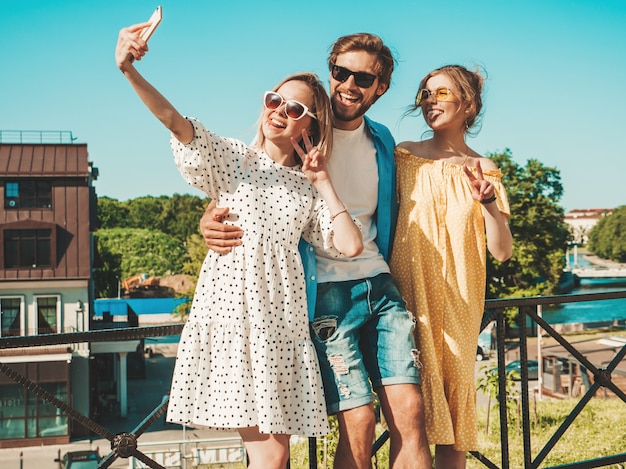 Grupo de tres jóvenes amigos con estilo en la calle. Hombre y dos chicas lindas vestidas con ropa casual de verano. Modelos sonrientes divirtiéndose en gafas de sol. Mujeres y hombres haciendo fotos selfie en teléfono inteligente