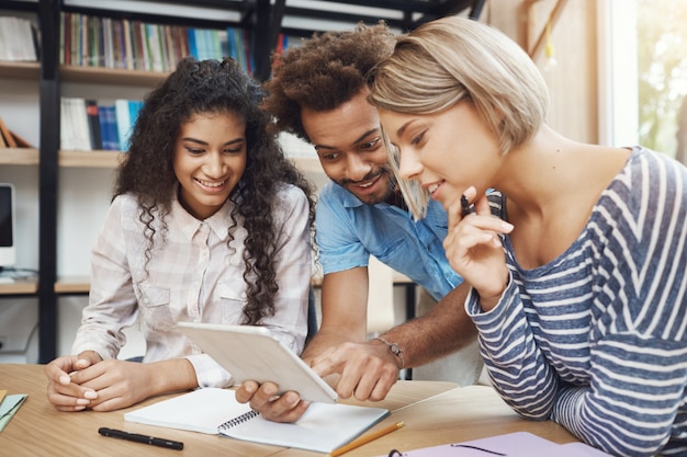 Grupo de tres jóvenes principiantes guapos sentados en un espacio ligero de coworking, hablando sobre proyectos futuros, mirando ejemplos de diseño en tabletas digitales. Amigos sonriendo, hablando de trabajo.