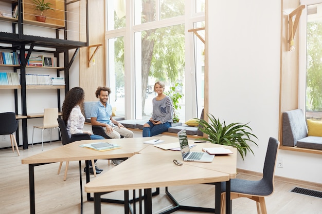 Grupo de tres jóvenes startups multiétnicas que trabajan juntas en el espacio de coworking, con un descanso de la lluvia de ideas. Jóvenes riendo, hablando, pasando un buen rato