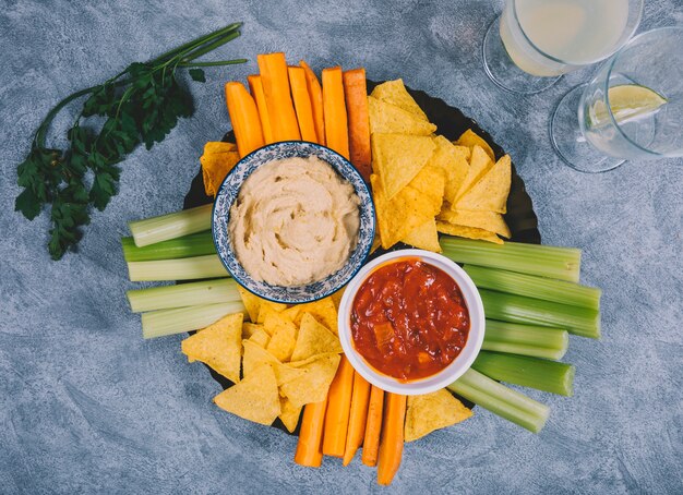 Guacamole y salsa de salsa en un bol con zanahoria; tallo de apio; jugo; Chips de cilantro y tortilla sobre fondo concreto