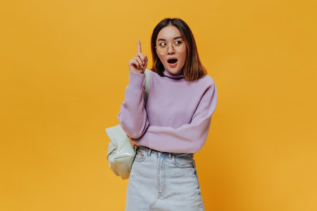 La guapa adolescente morena asiática con anteojos, suéter morado y falda de mezclilla apunta hacia arriba tiene una gran idea nueva y posa con mochila en el fondo naranja