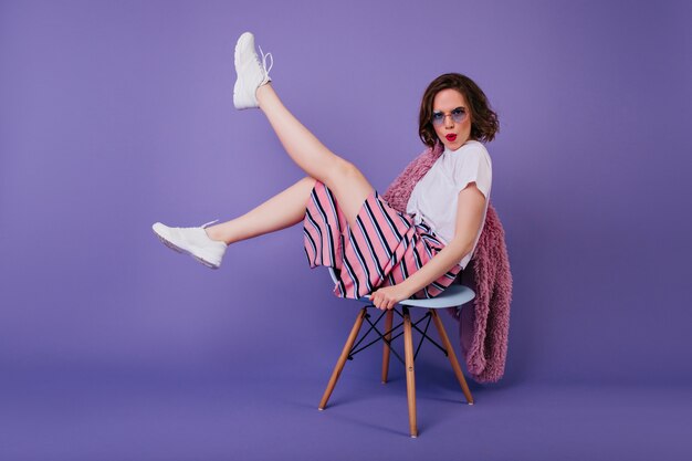 Guapa chica caucásica con maquillaje brillante sentado en una silla. Modelo de mujer relajada en zapatos blancos posando en la pared púrpura y agitando las piernas.