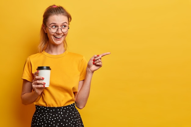 La guapa chica milenaria señala directamente en el espacio en blanco, invita a salir, muestra el camino a la publicidad, sostiene café para llevar, usa grandes anteojos redondos