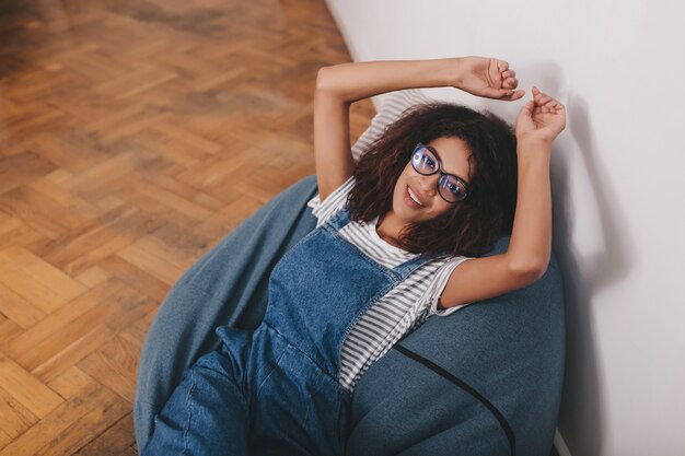 Guapa chica negra viste camisa a rayas y denim en general descansando en casa acostado sobre la almohada con las manos arriba