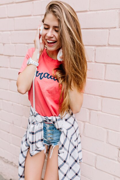 Guapa chica rubia con camisa rosa y pantalones cortos de mezclilla azul disfrutando de su música favorita en grandes auriculares blancos con los ojos cerrados