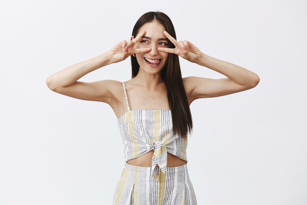 Guapa, despreocupada y juguetona chica con cabello oscuro en elegante blusa a juego con pantalones cortos, mostrando signos de victoria sobre los ojos, mirando a la izquierda y sonriendo con alegría, sacando la lengua