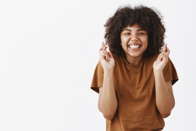 Guapa, fiel y optimista adolescente afroamericana con peinado afro cruzando los dedos para la buena suerte y sonriendo alegremente rezando por el sueño hecho realidad o pidiendo deseos